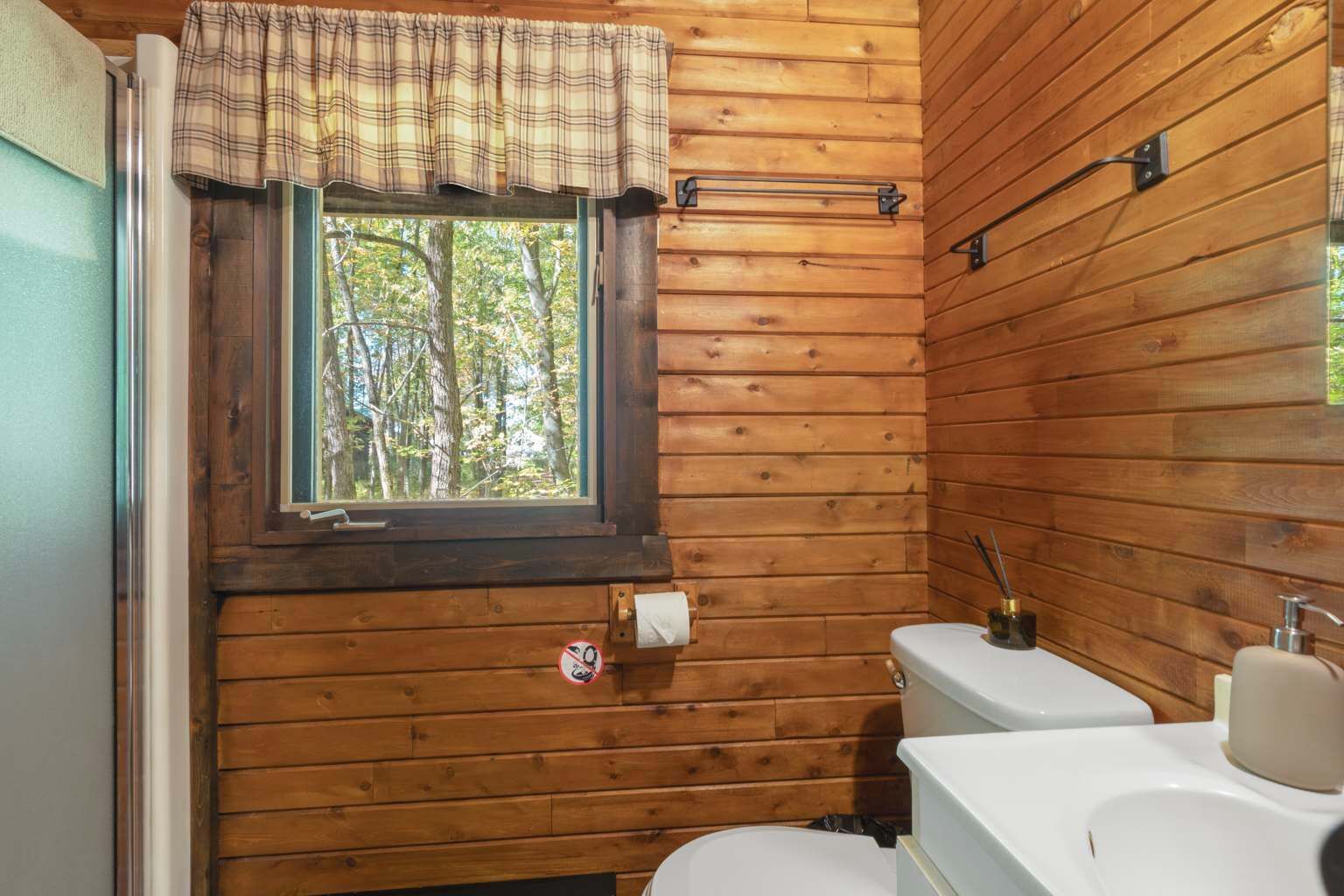 Baño de cabaña de madera con ventana, inodoro, lavabo y toallero.