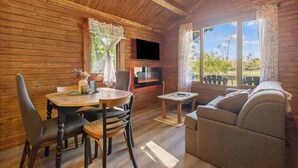 A cozy cabin interior with a wooden interior, a dining table with chairs, a sofa, a small table with a TV, and large windows with curtains.