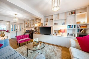 A spacious living room with a flat-screen TV, a piano, and a bookshelf filled with books and decorative items. - Charming Victorian London Home near Notting Hill – Perfect for Christmas (London)