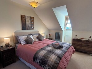 Ein Schlafzimmer mit schräg verlaufender Decke, einem Bett mit rosa Bettdecke, einem dunklen Kopfteil und einem Nachttisch mit Lampe.