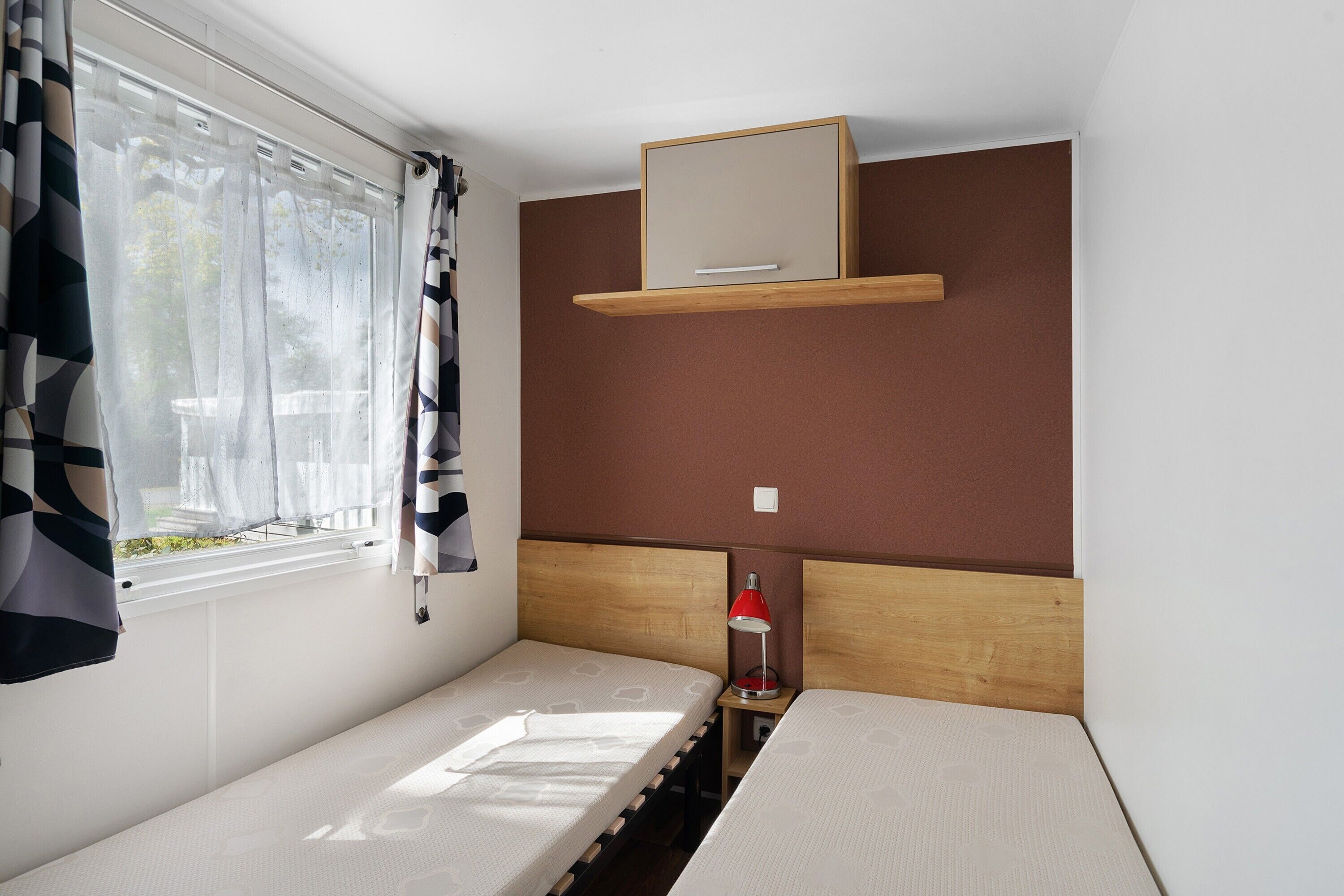 Una stanza modesta con due letti, una finestra con tende, un comodino con una lampada e una mensola in legno sopra i letti.