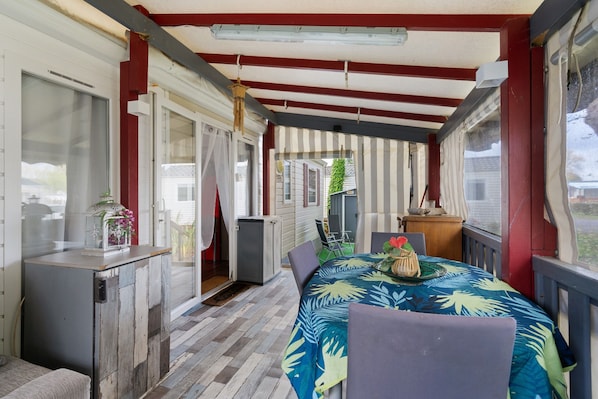 A covered outdoor dining area with a table set for two, a small refrigerator, and a window with curtains.