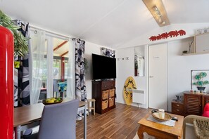 A modern living room with a flat-screen TV, wooden furniture, a dining area with a table and chairs, and a view through a glass door.