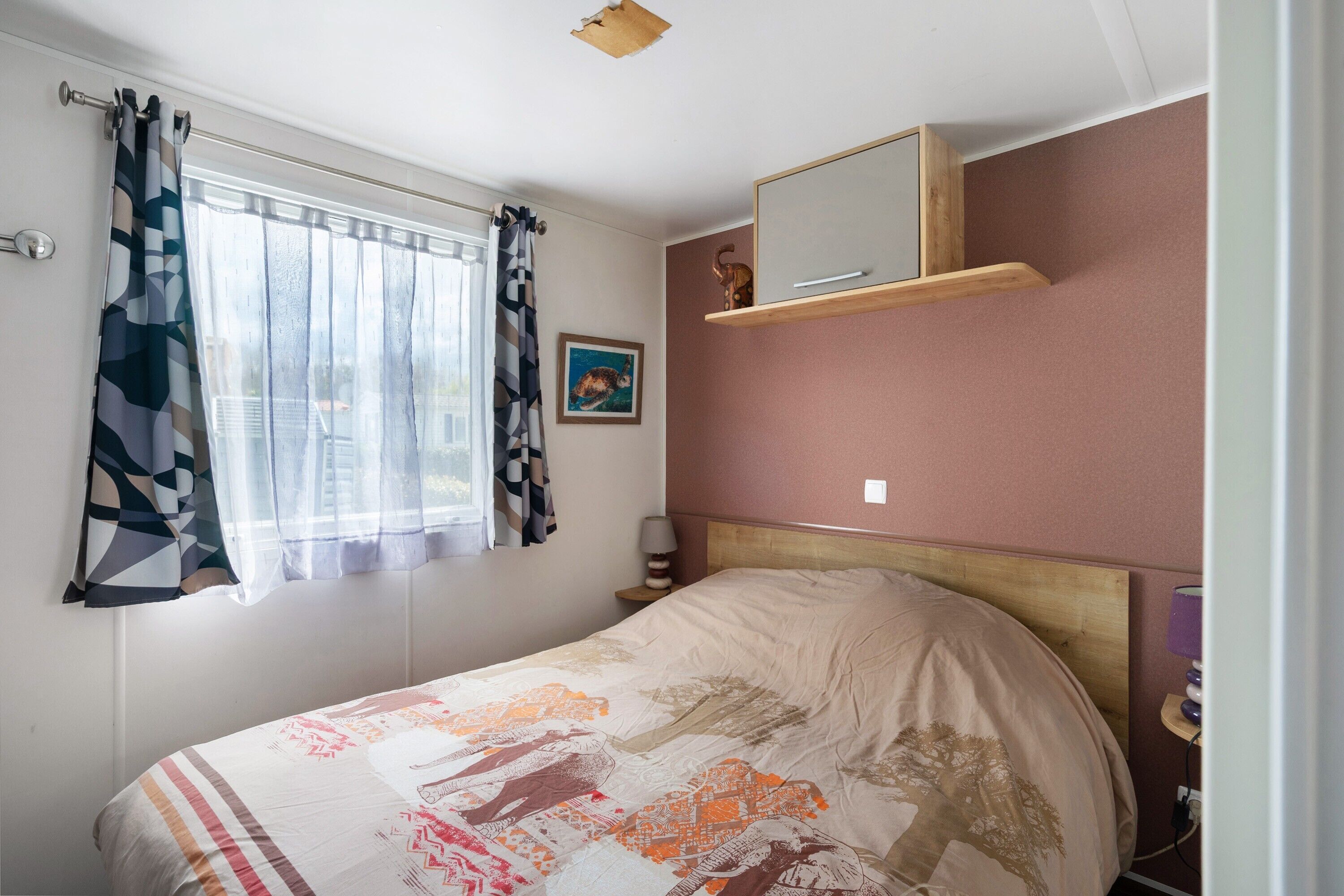 Ein Schlafzimmer mit Bett, Fenster mit Vorhängen, einer Nachttischlampe, einer an der Wand befestigten Ablage mit Spiegel und einem gerahmten Bild.