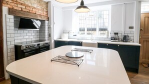 A modern kitchen with a large island, white cabinetry, a brick accent wall, and pendant lighting. - The Old Bakery (Banbury)