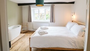 A bedroom with a wooden floor, a white bed, a window with curtains, a white bathtub, and a green pendant light. - The Old Bakery (Banbury)