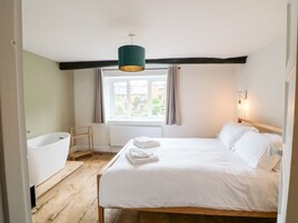 Un dormitorio con piso de madera, una cama blanca, una ventana con cortinas, una bañera blanca y una lámpara colgante verde.