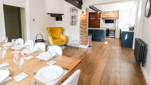 A dining area with a wooden table set for a meal, a yellow armchair, and a kitchen in the background. - The Old Bakery (Banbury)