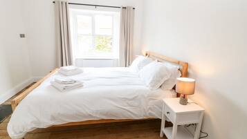 Cama bem arrumada com lençóis e toalhas brancas, abajur e janela com cortinas.