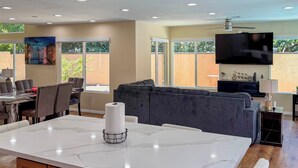 A modern kitchen with a marble island, a dining area with a table and chairs, a gray sofa, and a TV mounted on the wall. - Grill, Large Pool, Plush Patio Furniture, Umbrella (Rancho Cucamonga)