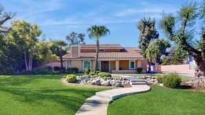A two-story house with a tiled roof, surrounded by a well-maintained lawn and a curved concrete pathway leading to the entrance. - Grill, Large Pool, Plush Patio Furniture, Umbrella (Rancho Cucamonga)
