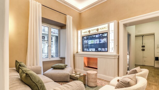 Een woonkamer met een open haard, een televisie erboven en een gezellige zithoek met een bank en fauteuils.