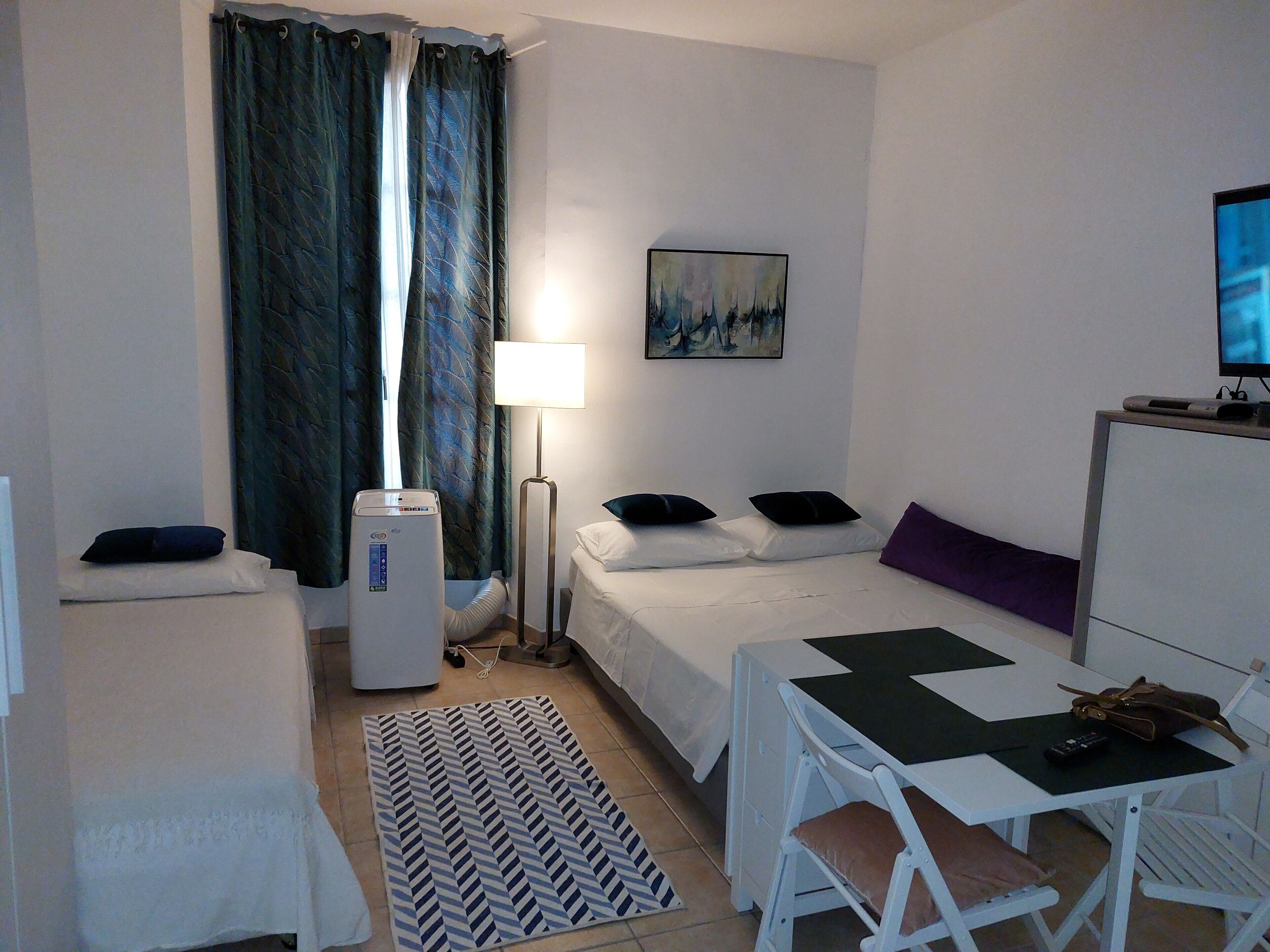 Quarto de hotel com cama, mesa de cabeceira, luminária, quadro, televisão e escrivaninha com cadeira.