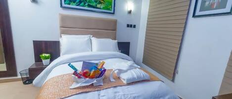 A neatly made bed with a white comforter and pillows, a bedside table with a plant, and a framed picture above the bed.