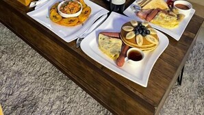 A breakfast setup on a wooden table with various dishes, a bottle of wine, and a person in white robe. - Morgan Hill Residence (Abuja)