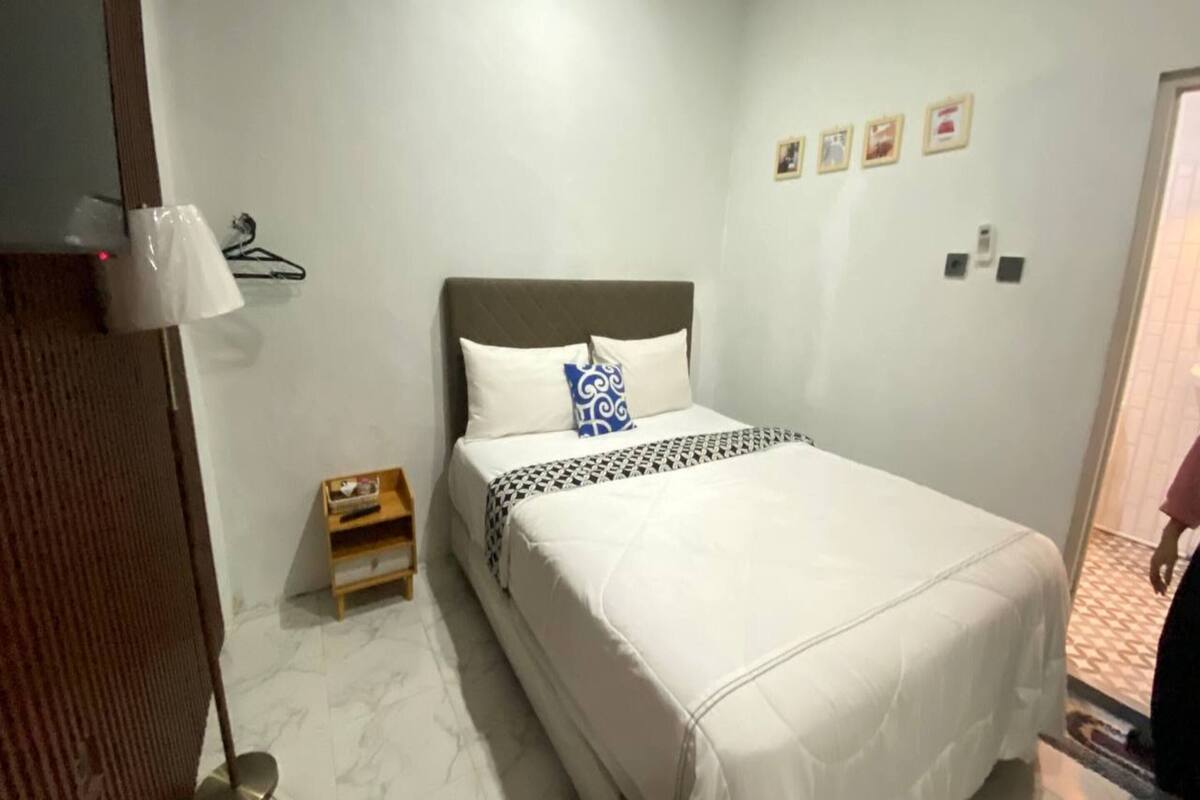 A neatly arranged hotel room with a bed, a small wooden nightstand, a wall-mounted lamp, and a door with a towel hanging on it.