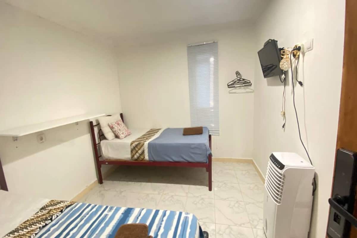 A small, well-lit room with a bed, a desk, a chair, a heater, and a window with blinds.