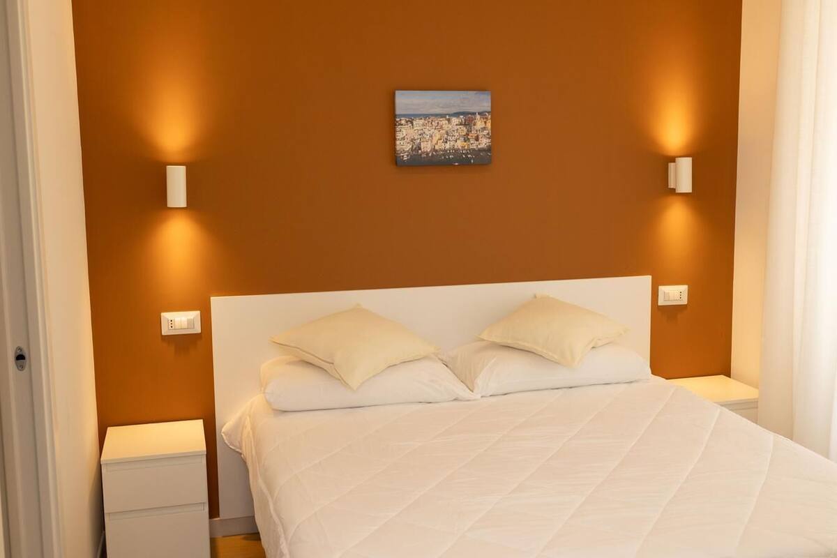 A bedroom with a white bed, two pillows, a nightstand, a wall-mounted light, and a painting above the bed.
