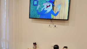Un televisor montado en la pared muestra un personaje de caricatura. Abajo, una mesita con una bandeja de objetos y una cafetera.