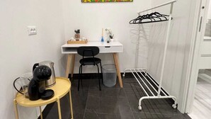 Una habitación pequeña y moderna con un escritorio blanco, una silla, una cafetera, una mesita, un tendedero y un cuadro abstracto de colores en la pared.