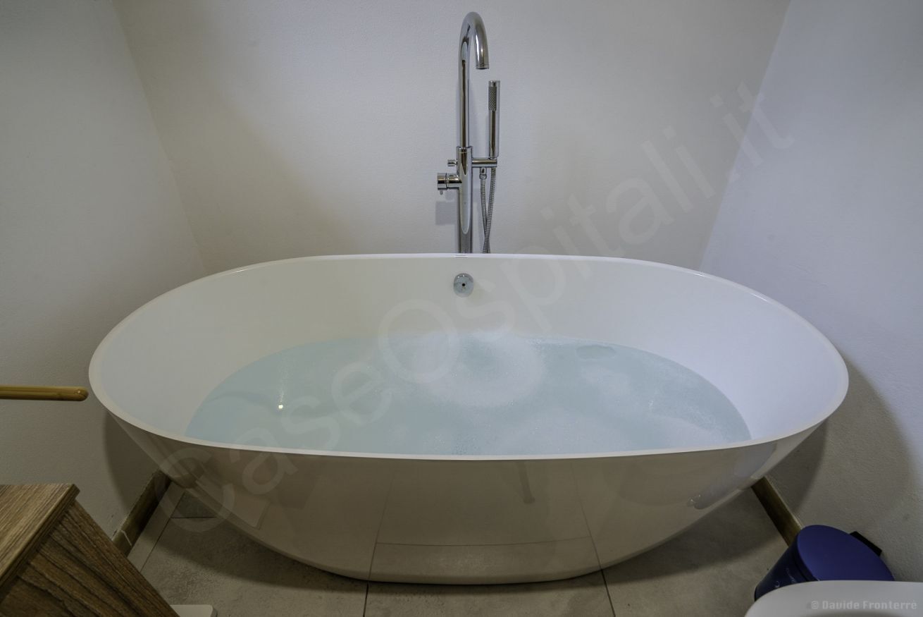 Eine moderne Badewanne mit Wasserhahn und Duschkopf, ein hölzerner Hocker und ein blaues Objekt auf dem Boden.