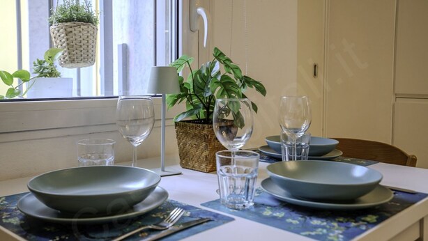 Une table à manger dressée avec des assiettes, des verres et des couverts, une plante en pot et une suspension florale.