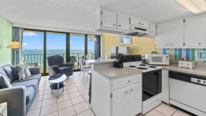 Une cuisine moderne avec des meubles blancs, une cuisinière, un four à micro-ondes et un grille-pain. On y trouve un coin repas avec une table et des chaises, ainsi qu’un canapé. La pièce est dotée de grandes fenêtres offrant une vue sur l’océan.