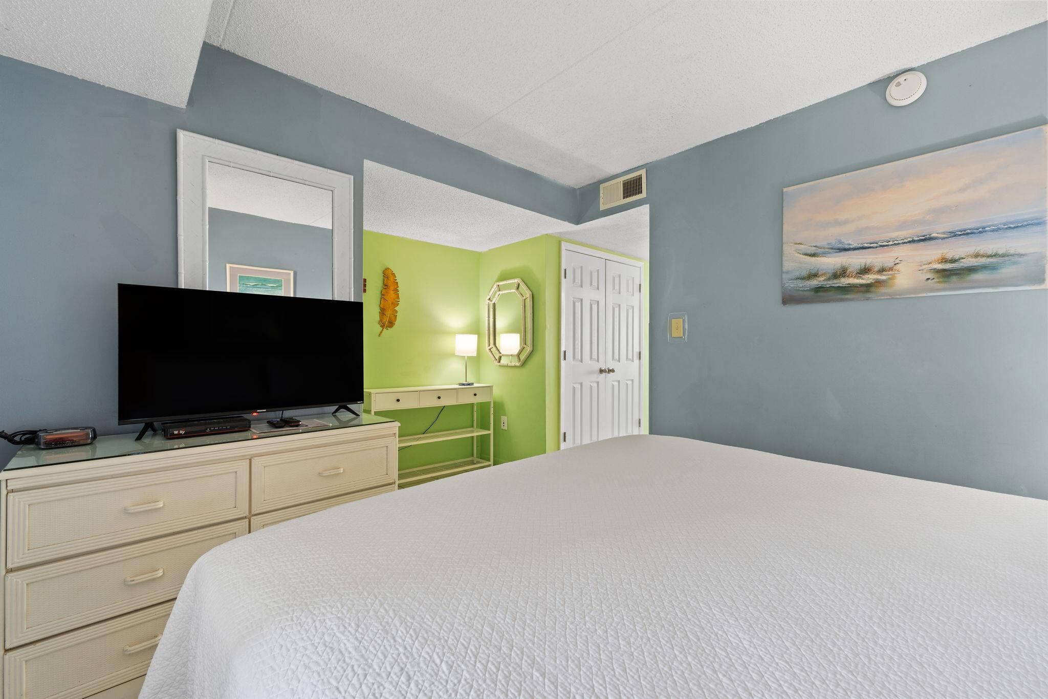Ein Schlafzimmer mit einem großen Bett, einem Flachbildfernseher auf einer Kommode, einer grünen Akzentwand, einer weißen Tür und einem Bild mit einer Strandansicht.