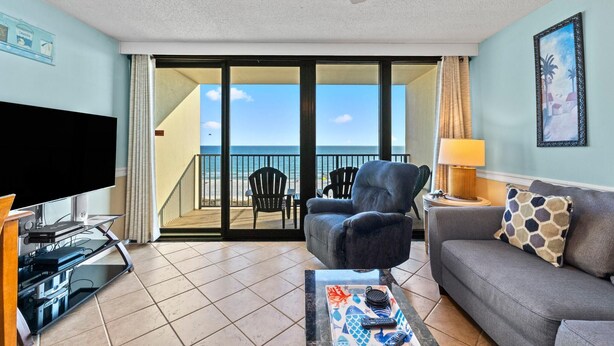 A living room with a TV, a sofa, a coffee table, and a balcony with a view of the ocean. - Island Winds West Unit 575 (Gulf Shores)
