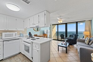 A modern kitchen with white cabinets, a microwave, and a stove. There is a dining area with a table and chairs, and a balcony with a view of the ocean. - Island Winds West Unit 575 (Gulf Shores)
