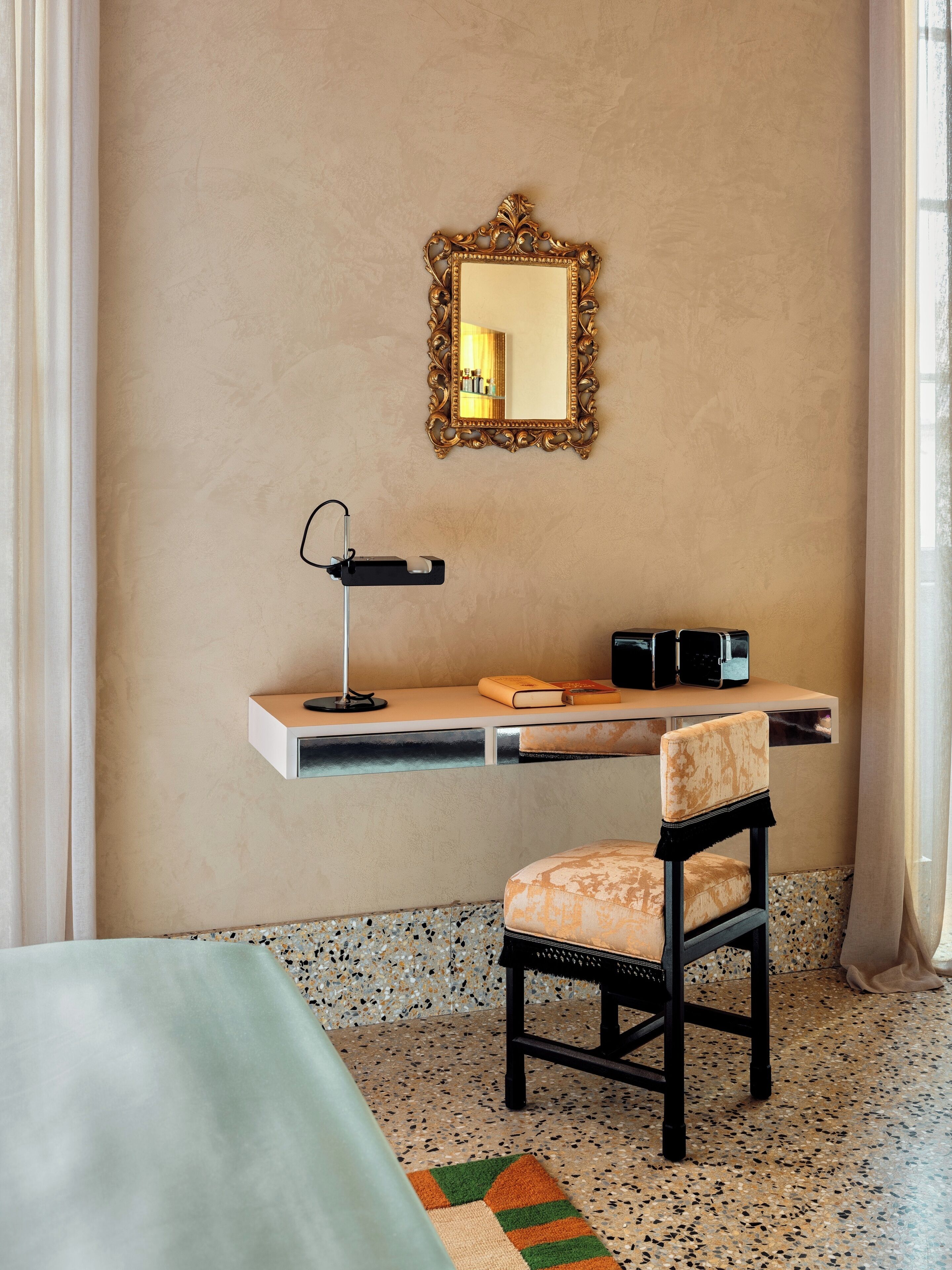 A room with a bed, a desk with a chair, a mirror, and a small shelf with books and a lamp.
