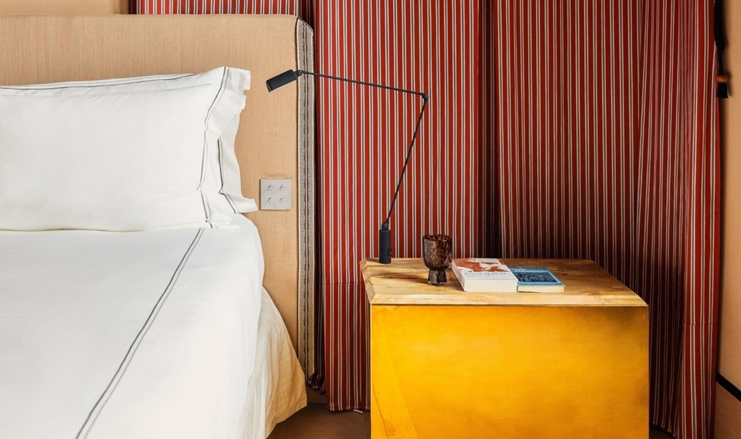 Hotellihuone, jossa on sänky, yöpöytä lampun kanssa, kirja ja koristeellinen kulho.