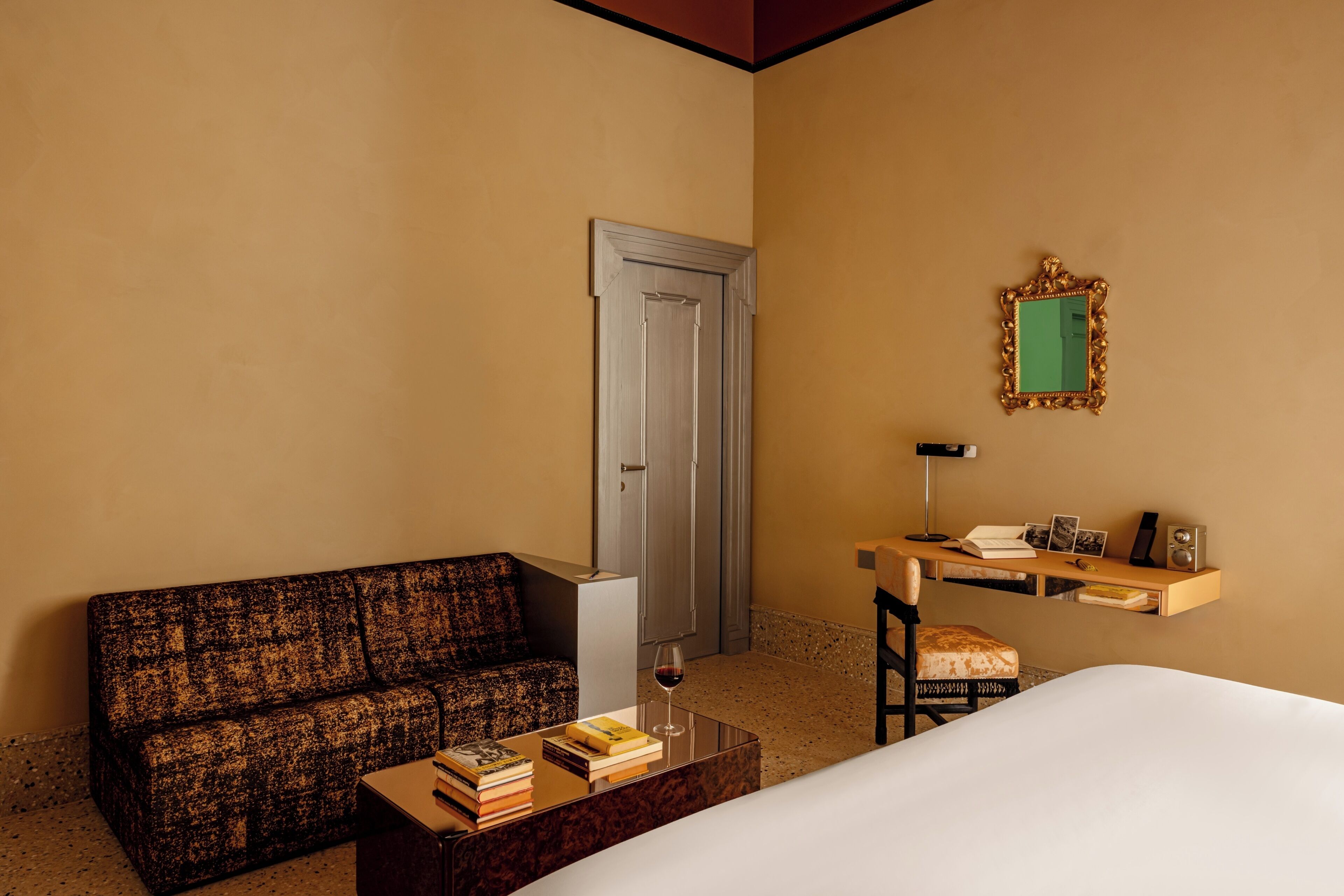 A hotel room with a sofa, a coffee table with books and a wine glass, a desk with a chair, a mirror, and a door.