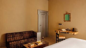 A hotel room with a sofa, a coffee table with books and a wine glass, a desk with a chair, a mirror, and a door.
