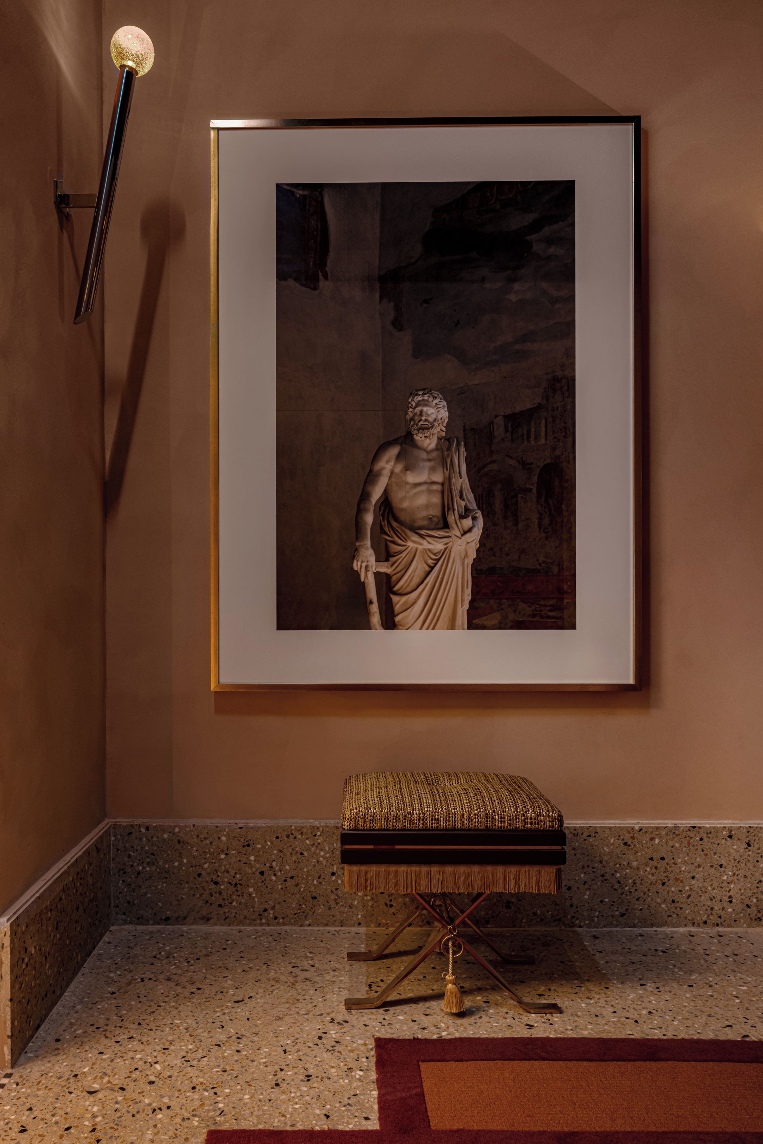 A framed black and white photograph of a classical statue on a wall, a small bench with a woven seat, and a terrazzo floor.