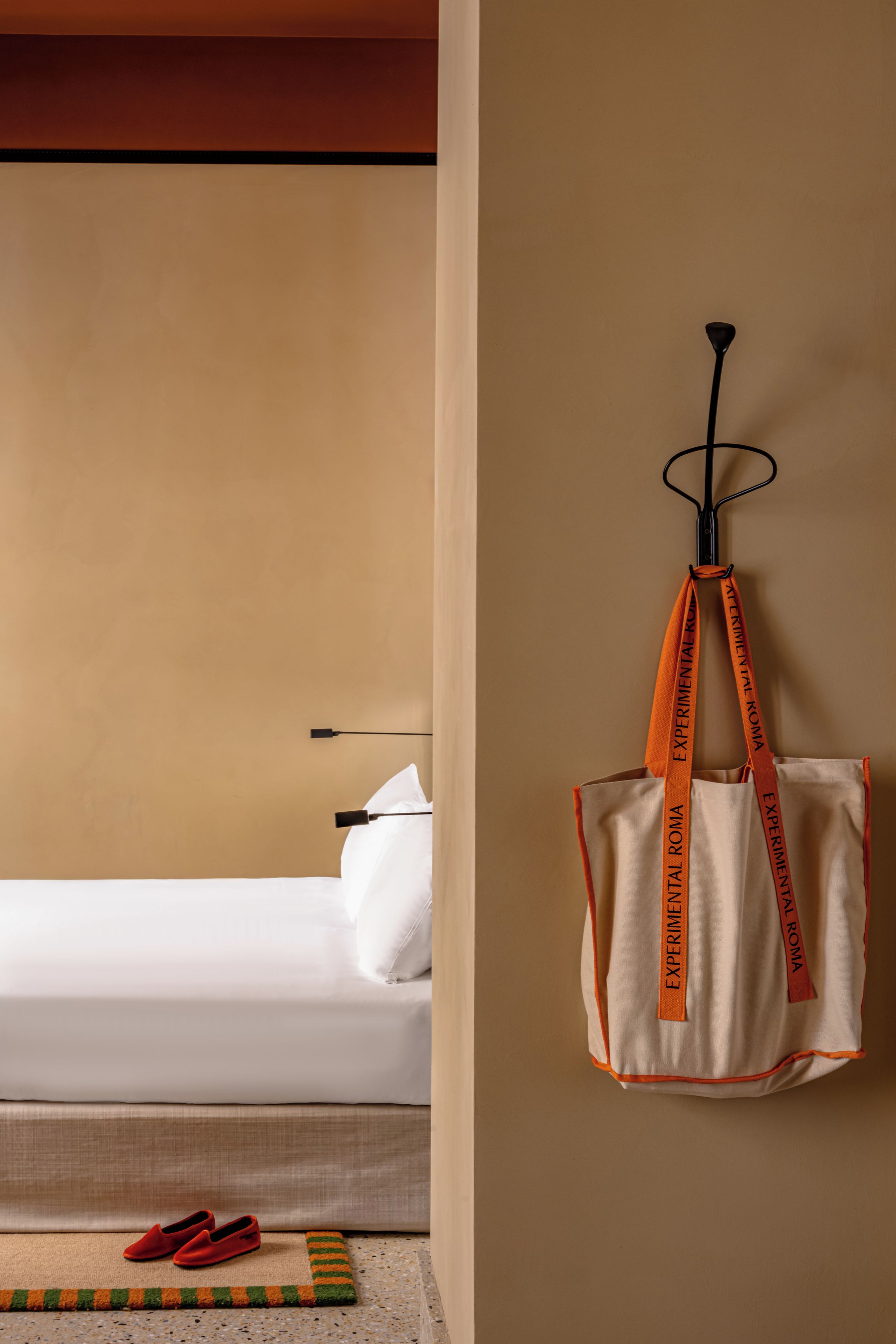 A hotel room with a bed, a wall-mounted bag rack, and a pair of slippers on the floor.