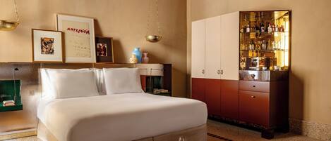 A hotel room with a bed, a minibar, a bookshelf, and a coffee table with books and a wine glass.