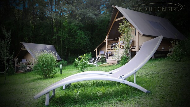 A modern outdoor lounge area with a curved white bench, surrounded by lush greenery and two wooden cabins with slanted roofs. - GLAMPING PIAN DELLE GINESTRE (Sassetta)
