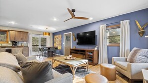 A modern living room with a sofa, coffee table, and ottoman. There is a TV, a ceiling fan, and a kitchen in the background. - Deadwood Pines Hideaway (Sturgis)
