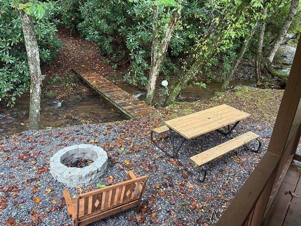 Un banc en bois, une table de pique-nique et un foyer, le tout installé dans un cadre naturel agrémenté d’arbres et d’un ruisseau.