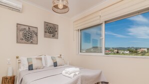 Een slaapkamer met een bed, twee decoratieve schilderijen, een raam met uitzicht en een nachtkastje met een lamp.