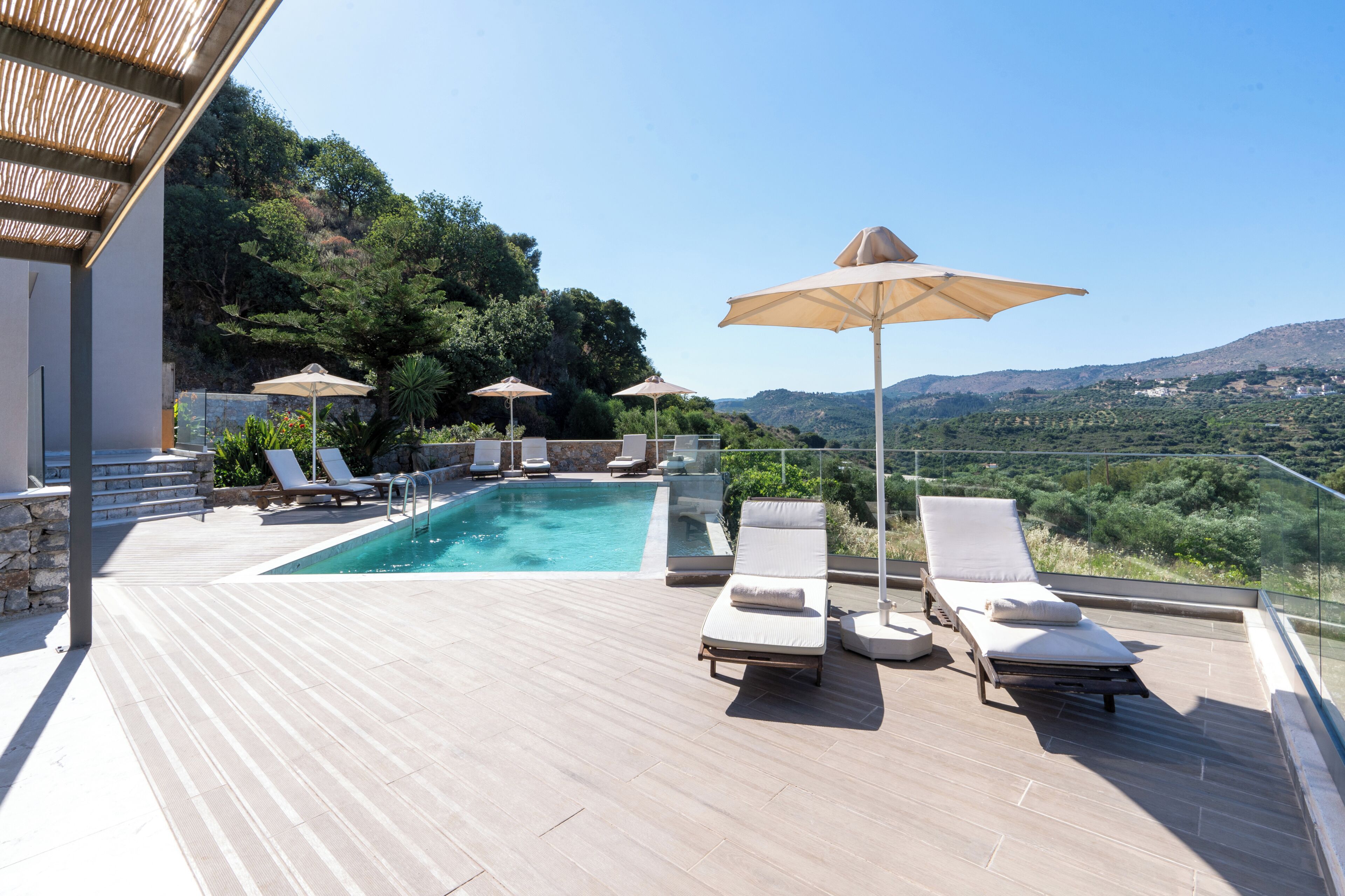 A rooftop pool area with lounge chairs and umbrellas, surrounded by a wooden deck and overlooking a mountainous landscape.