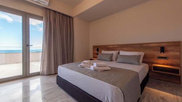 Quarto de hotel moderno com uma cama grande, cabeceira de madeira, mesa de cabeceira e vista para a praia através da porta de correr de vidro.