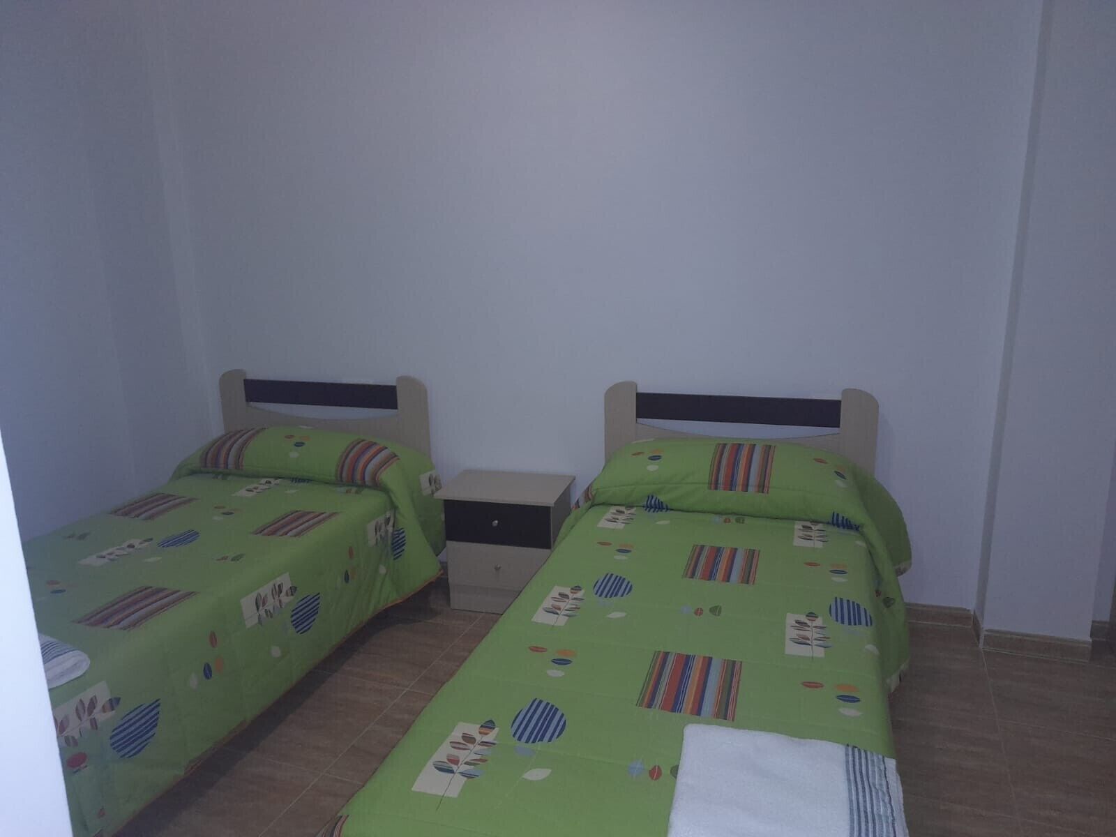 Een tweepersoonskamer met twee eenpersoonsbedden, elk met een groene spreidek en een houten hoofdbord. Er staat een klein nachtkastje met een lamp en een doos erop.