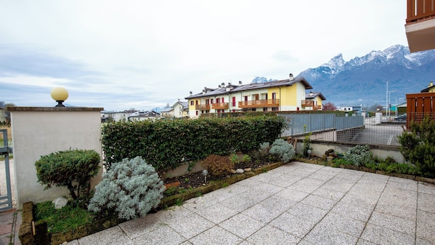 A residential area with a paved patio, a hedge, and a row of colorful houses with balconies, set against a backdrop of mountains. - Casa Shiva (Sedico)