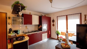 A modern kitchen with red cabinets, a gas stove, and a built-in oven. There is a dining area with a table and chairs, and a TV on a stand. - Casa Shiva (Sedico)