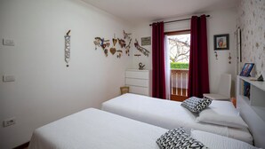 A bedroom with two beds, a dresser, a chair, and a balcony with red curtains. - Casa Shiva (Sedico)