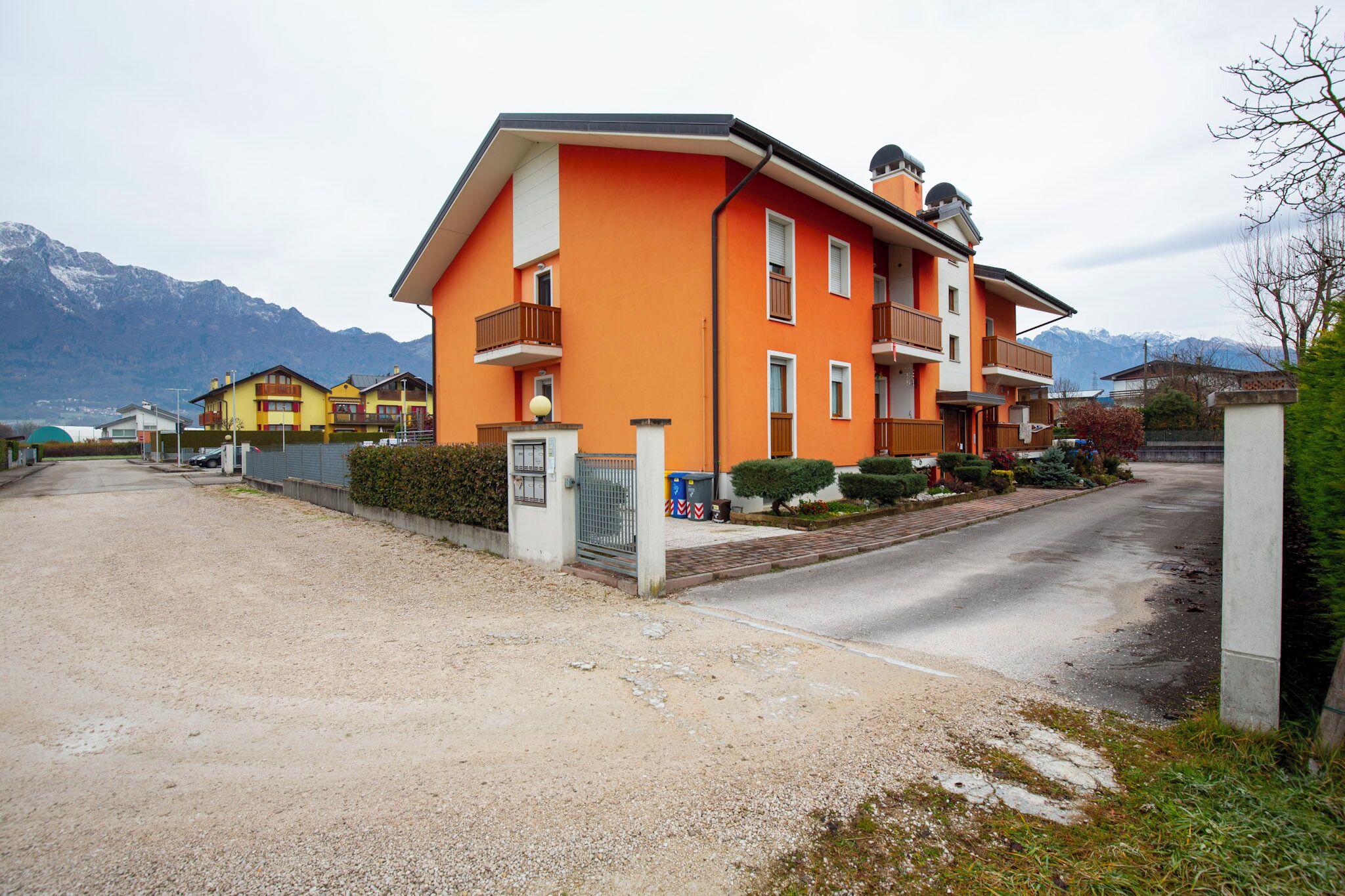 A multi-story building with balconies, a gravel driveway, and a mountain backdrop.