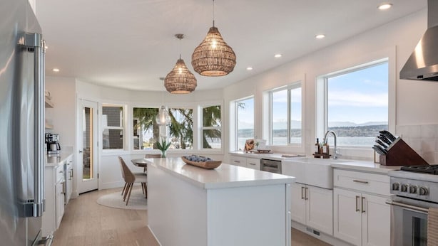 A modern kitchen with a central island, white cabinetry, stainless steel appliances, and large windows offering a view. - Pearl House Beach Retreat (Poulsbo)