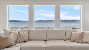 A beige sectional sofa with patterned cushions, positioned in front of three large windows offering a view of a calm body of water and distant land. - Pearl House Beach Retreat (Poulsbo)